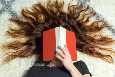 High angle view of woman with long hair covering face with book while relaxing on rug