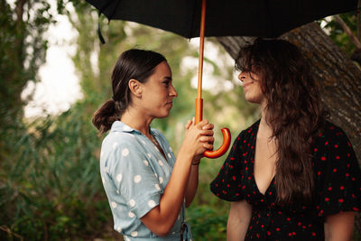 Young woman holding umbrella by friend while standing by tree trunk