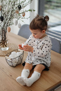 A little girl sits on a wooden table, near to her is a metal basket with easter eggs. 