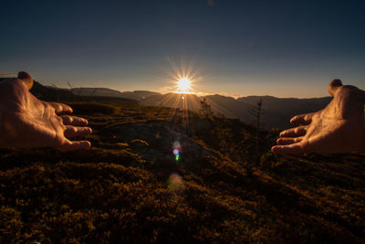 Cropped hands gesturing over landscape against sky during sunset