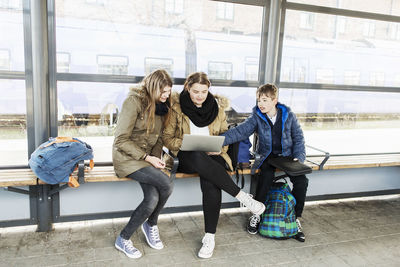 School students using laptop on bench at train station