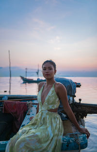 Portrait of young woman sitting at beach against sky during sunset