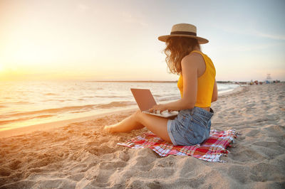 A slender young lady in denim shorts, a yellow tank top and a straw hat sits on a blanket 