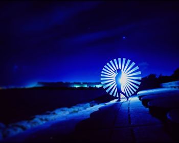 Illuminated ferris wheel by sea against sky at night