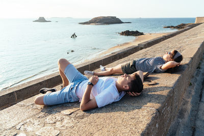 Friends lying on retaining wall against sea during sunset