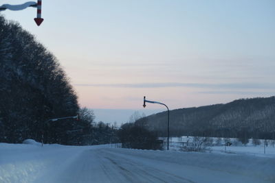 Snow covered road by trees against sky during sunset