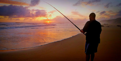 Man with fishing rod at beach against sky during sunset