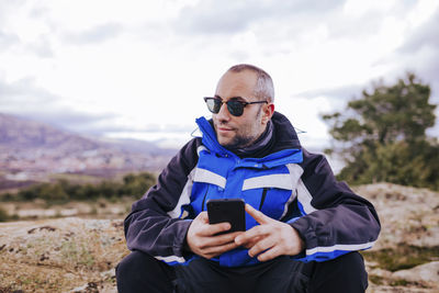 Man holding mobile phone while sitting on land against sky