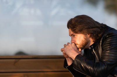 Pensive serious young bearded man wear black leather jacket sitting and thinking outdoors