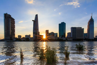 River by modern buildings against sky during sunset