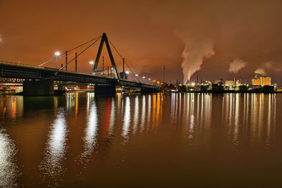 Illuminated highway bridge over a big river at night with industrial buildings in the background