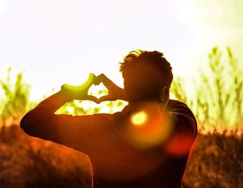 Rear view of man making heart shape with hands against sky during sunset