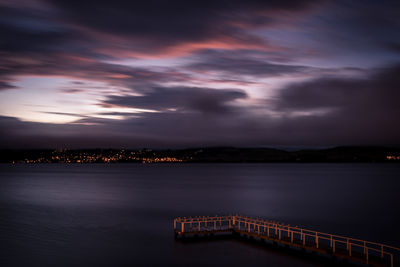 Illuminated city by sea against sky at sunset