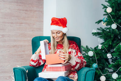 Cheerful woman wearing santa hat holding gift box while sitting by christmas tree at home