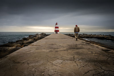 Rear view of man walking towards lighthouse at sea shore against cloudy sky