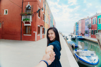 Smiling woman holding cropped hand by canal while standing against buildings in city