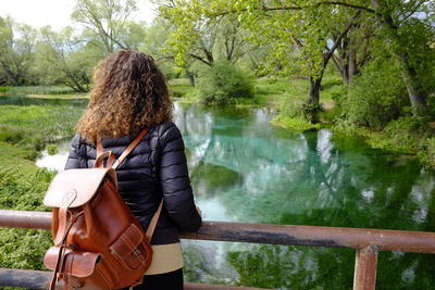 Rear view of woman with backpack standing on footbridge over river against trees