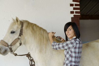 Portrait of smiling young woman combing horse hair while standing against wall