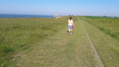 Rear view of woman walking on grass by sea against clear sky