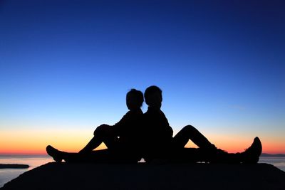 Silhouette couple sitting at beach against clear sky during sunset