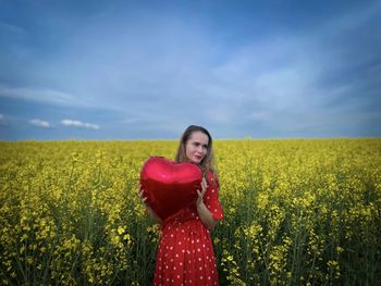 Woman in red dress standing in a field of canola flowers and holding a red heart balloon