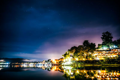 Illuminated buildings by lake against sky at night