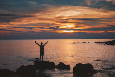 Silhouette man standing on rock by sea against sky during sunset