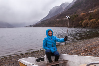 Woman in warm clothing taking selfie while sitting in boat at lakeshore