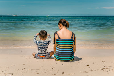 Rear view of mother with son sitting on beach against blue sky during sunny day