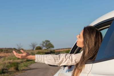 Excited young beautiful woman looking out the car window feeling the air with her arms raised 