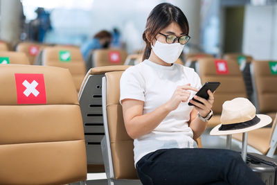 Woman wearing mask using phone sitting at airport