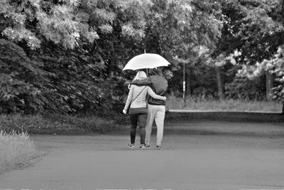Rear view of couple with umbrella walking on road against trees during monsoon
