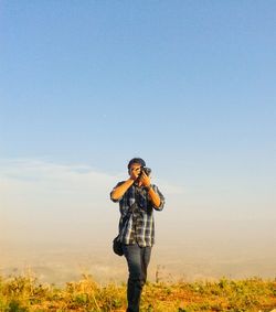 Young man photographing with digital camera on field against sky during sunset