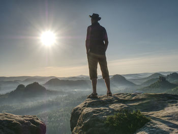 Tall man in shorts wearing cowboy hat shielding his face from sun, watching over large valley bellow