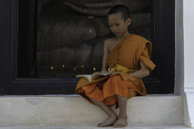 Boy wearing traditional clothing reading book while sitting on window sill