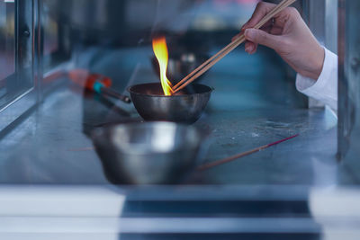 Cropped hand igniting incense sticks in bowl seen through glass