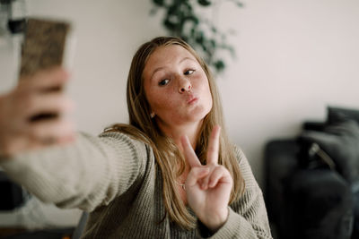 Teenage girl showing peace sign while taking selfie through mobile phone at home