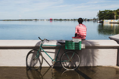 Rear view of man sitting on retaining wall by lake against sky