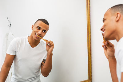 Smiling young man brushing teeth in front of mirror in bathroom