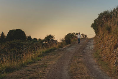Man walking on road amidst field against sky during sunset