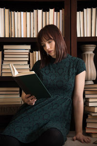 Young woman reading book while sitting against bookshelf in library