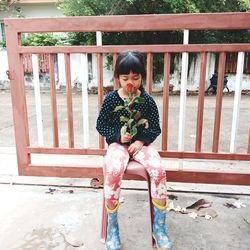 Full length of girl smelling flower while sitting on seat against gate