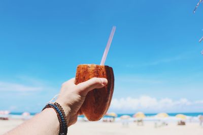 Cropped hand holding coconut against blue sky at beach during sunny day