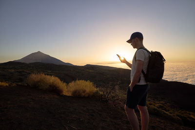 Hiker using mobile phone on hill above clouds at sunset. young man with backpack against landscape.
