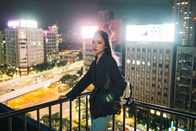 Portrait of woman standing by railing in city at night