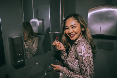 Portrait of smiling young woman applying lipstick in bathroom at nightclub