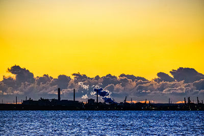 Silhouette factory by sea against sky during sunset