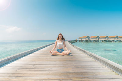 Woman meditating on pier over sea against blue sky