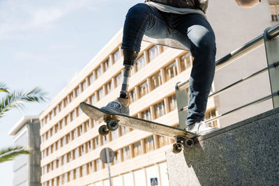 From below young guy with leg prosthesis jumping on skateboard above ground against modern building on sunny day