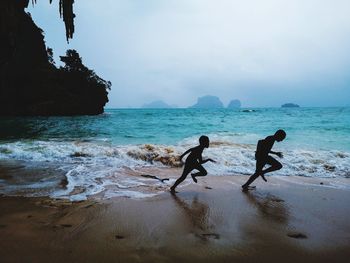 Silhouette boys running at beach against cloudy sky
